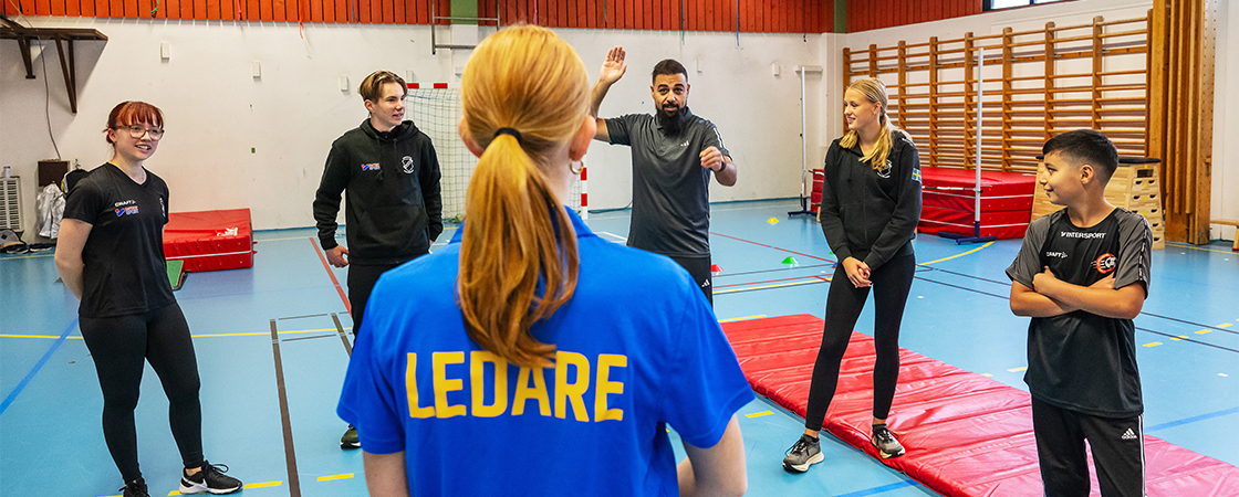 Fem ungdomar står i halvcirkel, en ledare med ryggen mot fotografen.